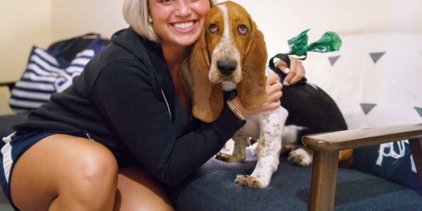 A smiling person embracing a basset hound on a couch.