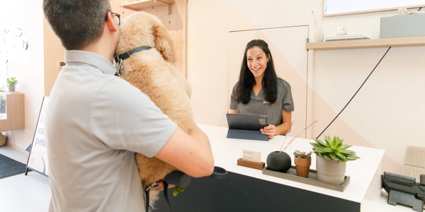 A man holding a dog at a reception while a woman behind the counter greets them with a smile.